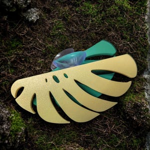 May include: A gold-colored, stylized monstera leaf-shaped object with three holes and a green base. A clear suction cup is attached. The object is set against a background of green moss and dark brown earth.
