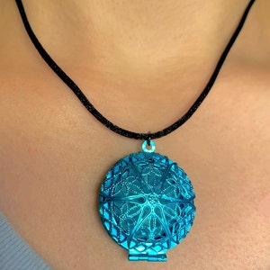 May include: A turquoise blue metal locket pendant with a delicate filigree design hangs from a black cord necklace.