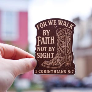 May include: A brown sticker featuring a cowboy boot design with intricate floral patterns. The sticker includes the text "FOR WE WALK BY FAITH, NOT BY SIGHT" and "2 CORINTHIANS 5:7" in white lettering. The sticker is held by a hand.