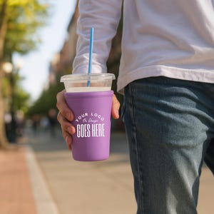 May include: A clear plastic cup with a blue straw and a purple drink sleeve. The sleeve has the text "YOUR LOGO GOES HERE" in white. The cup is held by a person wearing a white long-sleeve shirt and blue jeans.
