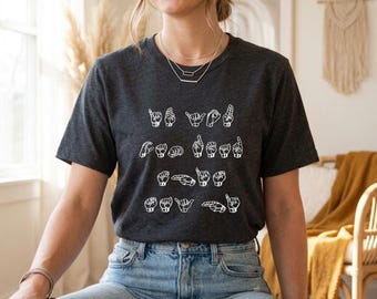 May include: A dark charcoal gray short-sleeve t-shirt featuring a white graphic print of American Sign Language hand signs arranged in four rows, spelling out a message across the chest. The shirt has a crew neckline and a standard fit.