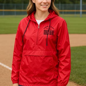 May include: A red hooded anorak jacket with a half-zip closure and a front pocket. The jacket has black drawstrings and the text "YOUR LOGO GOES HERE" printed in black. The model is wearing a white shirt underneath.