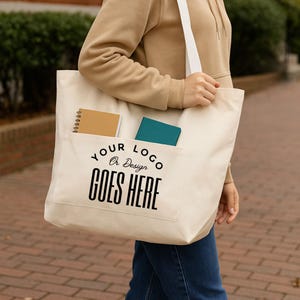 May include: A large off-white canvas tote bag with a front pocket featuring the black printed text YOUR LOGO OR DESIGN GOES HERE. The bag has white shoulder straps and contains a tan spiral notebook and a teal notebook inside the pocket.