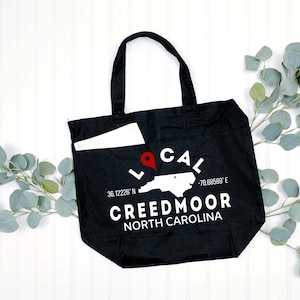 May include: Black canvas tote bag with a white graphic design featuring a map of North Carolina with a red pin, the words "LOCAL" and "CREEDMOOR NORTH CAROLINA", and the coordinates 36.12226° N -78.66589° E.