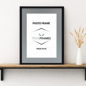 May include: A black rectangular photo frame with a grey mount. The white insert features the text PHOTO FRAME, PRIME FRAMES, and MADE IN UK. The frame is positioned on a wooden shelf next to a white vase containing dried wheat stalks.