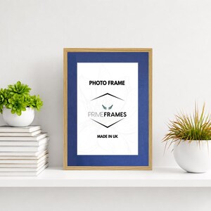 May include: A rectangular photo frame with a light wood finish and a deep blue mat board. The centre insert features the text PHOTO FRAME, the logo for PRIME FRAMES with a small wing icon, and the text MADE IN UK printed in black ink.