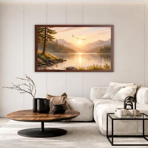 May include: A rectangular landscape wall art print in a dark brown wooden frame depicting a serene lake scene at sunrise. The image features calm water reflecting the golden sun, distant mountains, evergreen trees, and birds flying in the sky.