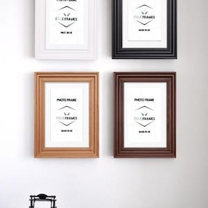 May include: Four rectangular photo frames in white, black, light brown, and dark brown hang on a white wall. Each frame has a white mat and the text "PHOTO FRAME" and "PRIME FRAMES MADE IN UK". Below are books and decor.