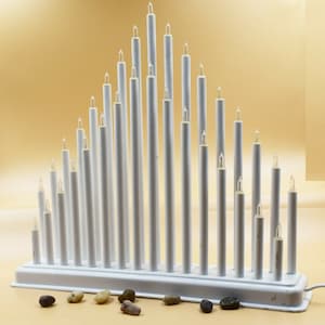 May include: White metal candle holder with a triangular shape and a row of lights. The holder is on a light brown surface with small stones scattered around it.