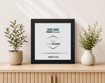 May include: A square black photo frame with a white insert featuring the text SQUARE PHOTO FRAMES, a logo with wings, the brand name PRIMEFRAMES, and the text MADE IN UK. The frame is positioned between two small green potted plants.