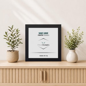 May include: A square black photo frame with a white insert featuring the text SQUARE PHOTO FRAMES, a logo with wings, the brand name PRIMEFRAMES, and the text MADE IN UK. The frame is positioned between two small green potted plants.
