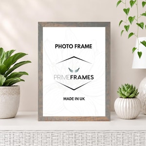 May include: A rectangular photo frame with a weathered gray wood finish. The white insert features the black text PHOTO FRAME, the brand name PRIMEFRAMES with a small wing icon, and the text MADE IN UK, all centered within a thin black diamond outline.