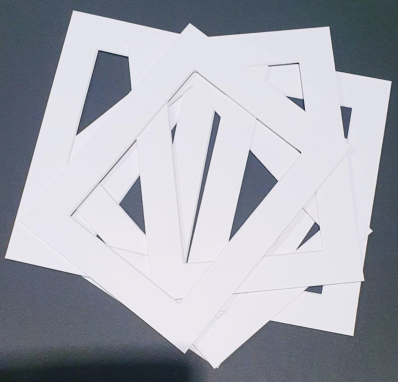 May include: A stack of white picture frame mats with square openings. The mats are made of thick, white cardstock and are arranged in a slightly overlapping pattern, revealing the dark surface beneath.