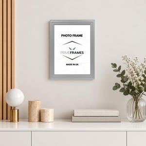 May include: A rectangular silver photo frame with a beveled edge. The insert features the text PHOTO FRAME, the logo PRIME FRAMES with a small wing icon, and the text MADE IN UK. The frame is displayed vertically against a plain wall surface.