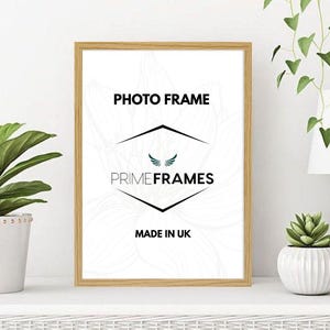 May include: A rectangular photo frame with a light oak wood finish. The white insert features the black text PHOTO FRAME, the brand name PRIMEFRAMES with a small wing icon, and the text MADE IN UK centred within a black diamond outline.