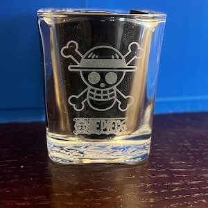 May include: A clear glass shot glass featuring an etched design of the One Piece skull and crossbones logo with a straw hat on the skull. Below the skull, the text ONE PIECE is etched into the glass surface. The glass has a square base.