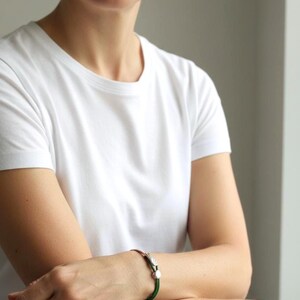 May include: A silver and copper bracelet with a green cord. The bracelet features a decorative feather design and a small, round clasp. The person in the image is wearing a white t-shirt.