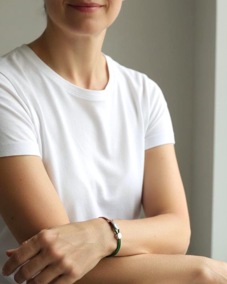 May include: A silver and copper bracelet with a green cord. The bracelet features a decorative feather design and a small, round clasp. The person in the image is wearing a white t-shirt.