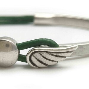 May include: A silver-toned bracelet with a green leather strap. The bracelet features a wing-shaped clasp and a circular metal accent. The design is minimalist and modern, suitable for everyday wear.