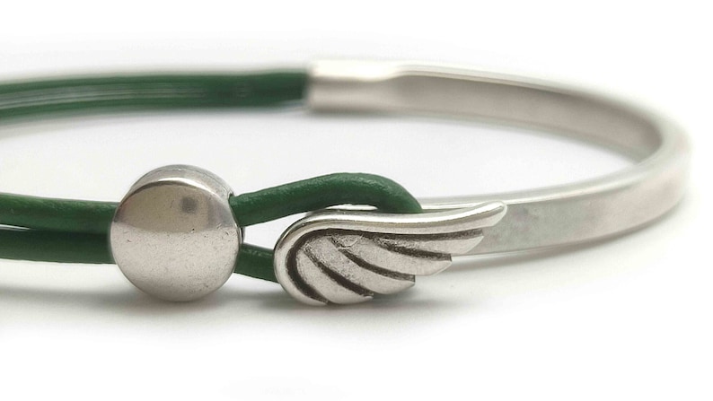 May include: A silver-toned bracelet with a green leather strap. The bracelet features a wing-shaped clasp and a circular metal accent. The design is minimalist and modern, suitable for everyday wear.