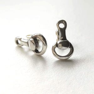 Può includere: Un paio di gemelli color argento, ciascuno a forma di morso di cavallo. I gemelli hanno una superficie lucida e riflettente e sono posti su uno sfondo bianco. Il design comprende un anello e una barra.