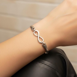 Op de afbeelding: Een zilverkleurige armband met een centraal schakel in de vorm van een oneindigheidssymbool. De armband is opgebouwd uit gladde, gepolijste metalen segmenten die verbonden zijn met de oneindigheidslus voor een strak en doorlopend ontwerp.