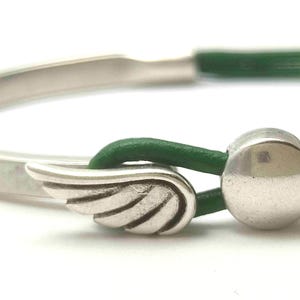 May include: Silver bracelet with a green leather cord. The bracelet features a wing charm and a silver ball clasp. The design is simple and elegant, suitable for everyday wear. The bracelet is a stylish accessory.