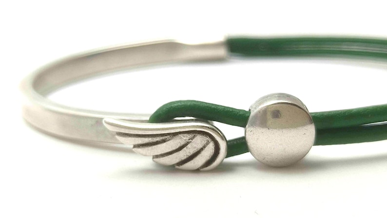 May include: Silver bracelet with a green leather cord. The bracelet features a wing charm and a silver ball clasp. The design is simple and elegant, suitable for everyday wear. The bracelet is a stylish accessory.
