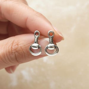 Pode incluir: Dois pequenos berloques de metal prateado em forma de bridão de cavalo. Cada berloque apresenta uma barra horizontal central ligada a dois anéis circulares e um pequeno laço no topo para fixação, com um acabamento polido e reflexivo.