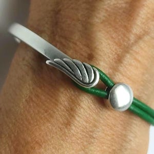 May include: A silver-toned bracelet with a wing-shaped accent and a green leather band. The bracelet features a curved bar and a round clasp. The leather band is approximately 0.25 inches wide.