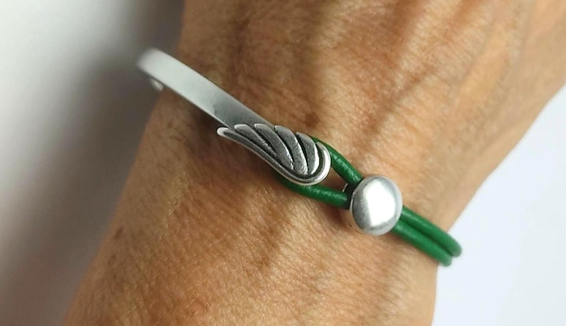 May include: A silver-toned bracelet with a wing-shaped accent and a green leather band. The bracelet features a curved bar and a round clasp. The leather band is approximately 0.25 inches wide.
