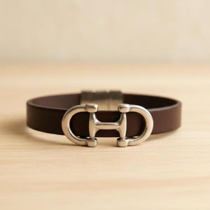 以下が含まれることがあります： ダークブラウンのレザーブレスレット。中央には、馬のハミを模したシルバーカラーの金属製クラスプが付いています。滑らかなレザーバンドは、機能的な留め具と装飾的な要素の両方を兼ね備えた金属製の金具によって固定されています。