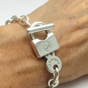 Puede incluir: Un brazalete de cadena de plata con un dije de candado. El candado tiene el número "24" grabado en él. El dije también tiene el texto "MADE IN EEC" grabado en él.