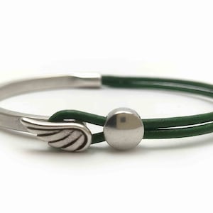 May include: A silver-toned bracelet with a green leather band. The bracelet features a wing charm and a silver ball clasp. The design is simple and elegant, suitable for everyday wear. The bracelet is shown against a white background.