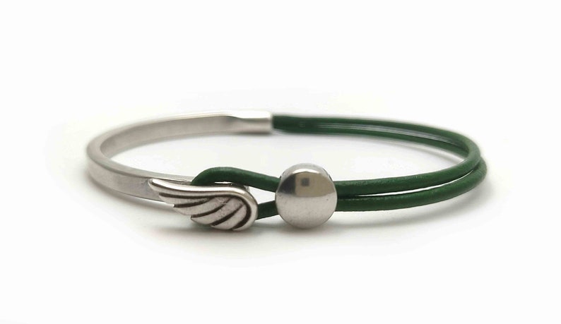 May include: A silver-toned bracelet with a green leather band. The bracelet features a wing charm and a silver ball clasp. The design is simple and elegant, suitable for everyday wear. The bracelet is shown against a white background.