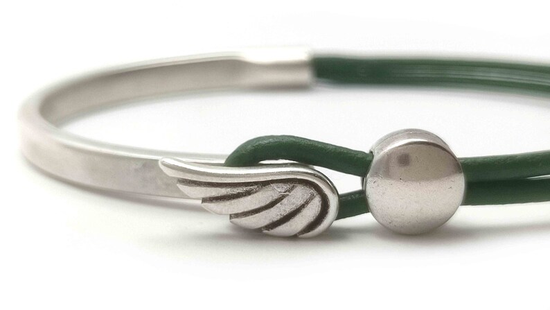 May include: A silver bracelet with a green leather band. The bracelet features a wing charm and a round silver bead. The design is simple and elegant, suitable for everyday wear.