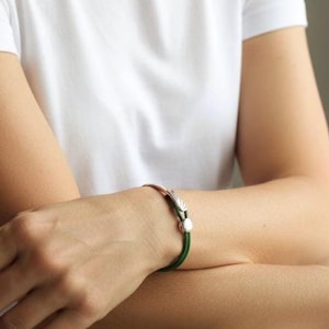 May include: A copper-toned bracelet with a green cord and silver accents. The bracelet features a decorative silver clasp and a small, round silver bead. The bracelet is worn on a wrist, with a white shirt in the background.