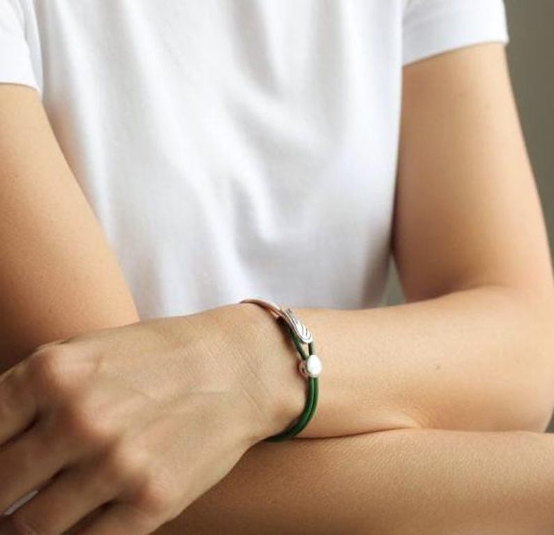 May include: A copper-toned bracelet with a green cord and silver accents. The bracelet features a decorative silver clasp and a small, round silver bead. The bracelet is worn on a wrist, with a white shirt in the background.