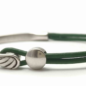 May include: A silver-toned bracelet with a green leather band. The bracelet features a wing charm and a circular clasp. The green leather band is doubled, and the bracelet is set against a white background.