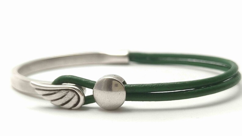 May include: A silver-toned bracelet with a green leather band. The bracelet features a wing charm and a circular clasp. The green leather band is doubled, and the bracelet is set against a white background.