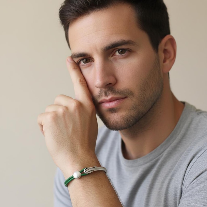 May include: A silver bracelet with a green band and a white pearl. The bracelet features a wing design on the clasp. The bracelet is worn on a person's wrist.
