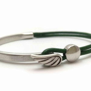 May include: A silver-toned bracelet with a green leather band. The bracelet features a wing charm and a circular clasp. The design is minimalist and modern, suitable for everyday wear. The bracelet's overall length is approximately 7 inches.