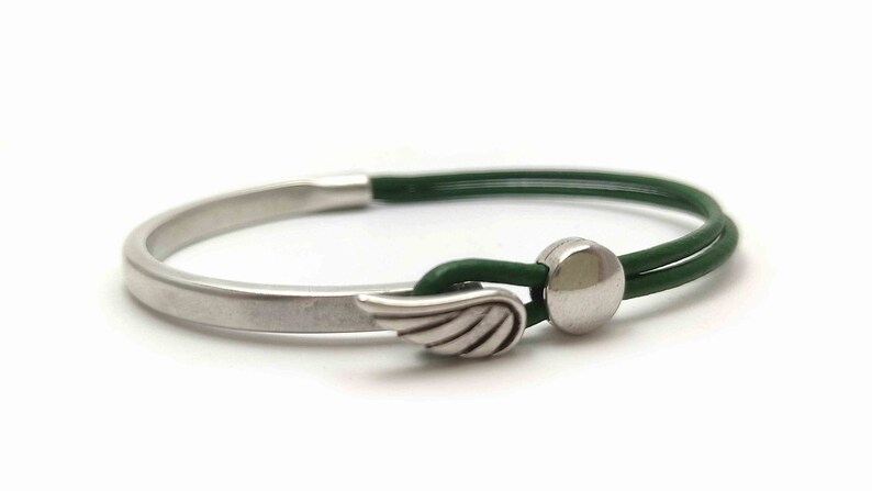 May include: A silver-toned bracelet with a green leather band. The bracelet features a wing charm and a circular clasp. The design is minimalist and modern, suitable for everyday wear. The bracelet's overall length is approximately 7 inches.