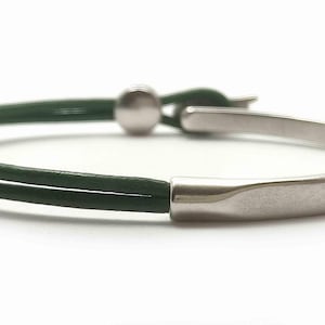 May include: A minimalist bracelet featuring a dark green leather band and a silver-toned metal clasp. The bracelet has a curved, rectangular metal piece and a small, round ball for closure. The design is simple and modern.