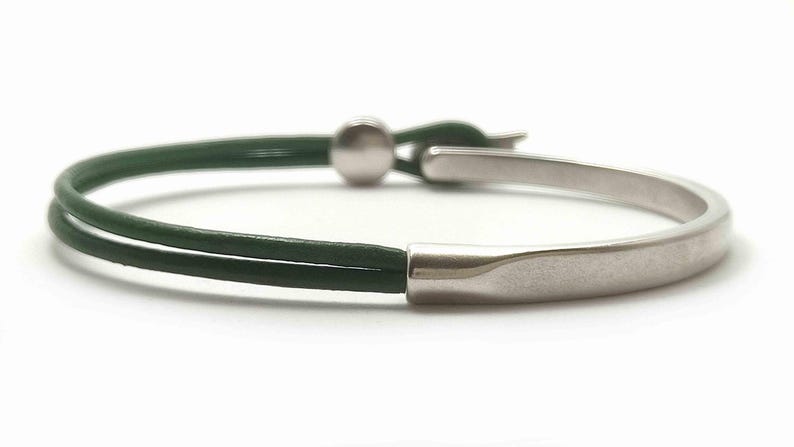 May include: A minimalist bracelet featuring a dark green leather band and a silver-toned metal clasp. The bracelet has a curved, rectangular metal piece and a small, round ball for closure. The design is simple and modern.