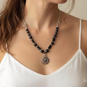 Peut inclure: Un collier composé d'un pendentif en forme de fossile d'ammonite en argent, suspendu à une chaîne de perles de lave noire poreuse et de perles cylindriques noires, agrémenté de petites entretoises argentées et d'un fermoir à bascule.