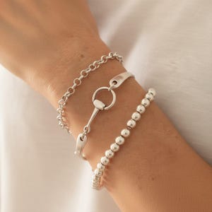 May include: A set of three silver-toned bracelets. One bracelet features a delicate round link chain, the second is a rigid cuff with a central circular horse-bit style clasp, and the third is composed of a single strand of polished silver beads.