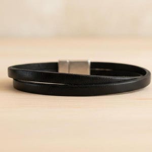 Peut inclure: Un bracelet en cuir noir à double tour doté d'une finition lisse et mate ainsi que d'un fermoir magnétique rectangulaire en métal argenté. Le bracelet est composé de deux cordons en cuir fins et parallèles qui se chevauchent.