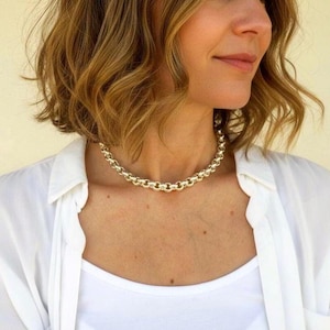 Può includere: Una collana a catena color oro con un design a maglie arrotondate. La collana è indossata sopra un top bianco e una camicia bianca abbottonata. La collana è uno stile choker.