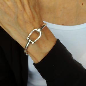 May include: A polished silver-tone bangle bracelet featuring a horseshoe-shaped clasp mechanism. The bracelet has a smooth, reflective metallic finish and a rounded band design that secures with a central U-shaped link and two small spherical accents.